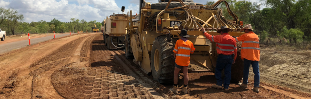 Three construction workers standing against machinery on dirt covered road